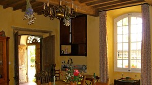 Ein rustikaler Essraum mit einem Holztisch, Stühlen und einer Deckenleuchte. Ein Fenster mit Vorhängen und eine Tür, die in einen anderen Raum führt.