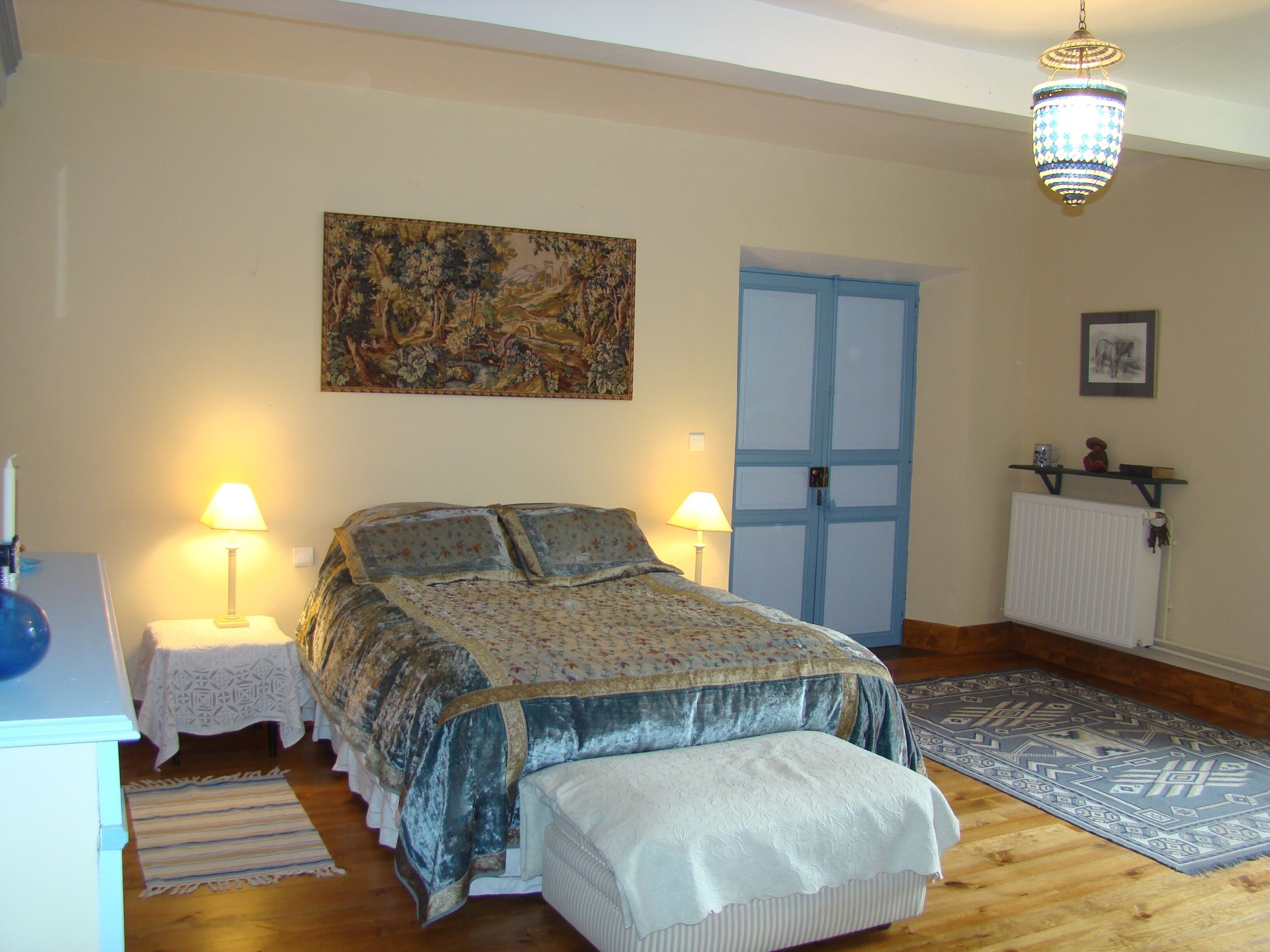 A bedroom with a bed, two bedside lamps, a painting on the wall, a blue door, a radiator, and a patterned rug.