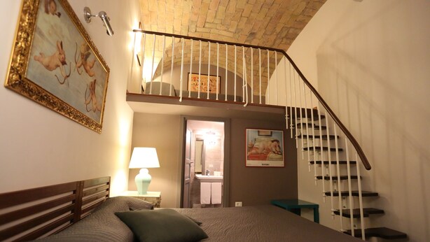 A bedroom with a bed, a nightstand, a lamp, a painting, a staircase, and a bathroom visible through an open door.