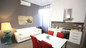 A modern kitchen with white cabinets, a stainless steel stove, and a refrigerator. There is a dining area with a white table and red chairs. A white sofa and a lamp are in the living area.