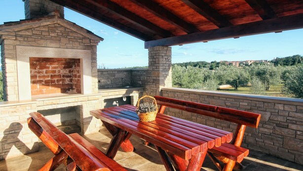 Un patio al aire libre con chimenea de piedra, bancos de madera y vistas a olivos y edificios lejanos.