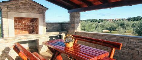 An outdoor patio with a stone fireplace, wooden benches, and a view of olive trees and distant buildings.