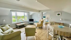 A spacious living room with a large window, a television, a dining table, and a cozy seating area. - Clemens Beach House, Sea Views, Private Lane, Crantock, Sleeps 4. (Crantock, Cornwall)