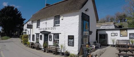 A white building with a black roof, labeled "Old Albion Inn." There are outdoor seating areas with benches and tables, and a signboard with information.