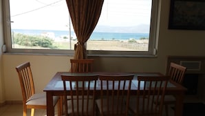 A dining area with a table and chairs, a window with a view of the sea and distant buildings, and a curtain partially drawn.