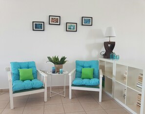 Ein Zimmer mit zwei blauen Sesseln mit grünen Kissen, einem kleinen Tisch mit einer Pflanze, einem weißen Schrank mit Büchern und vier gerahmten Kunstwerken an der Wand.