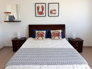 Ein ordentlich bezogenes Bett mit einem gemusterten Überwurf, zwei Kissen mit Krappendruck und einem Holzkopfteil. Ein Nachttisch mit Lampe steht daneben.