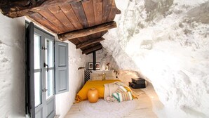 Une petite pièce rustique avec un plafond en bois, un lit recouvert d’une couverture jaune, une petite table avec une lampe et une valise.