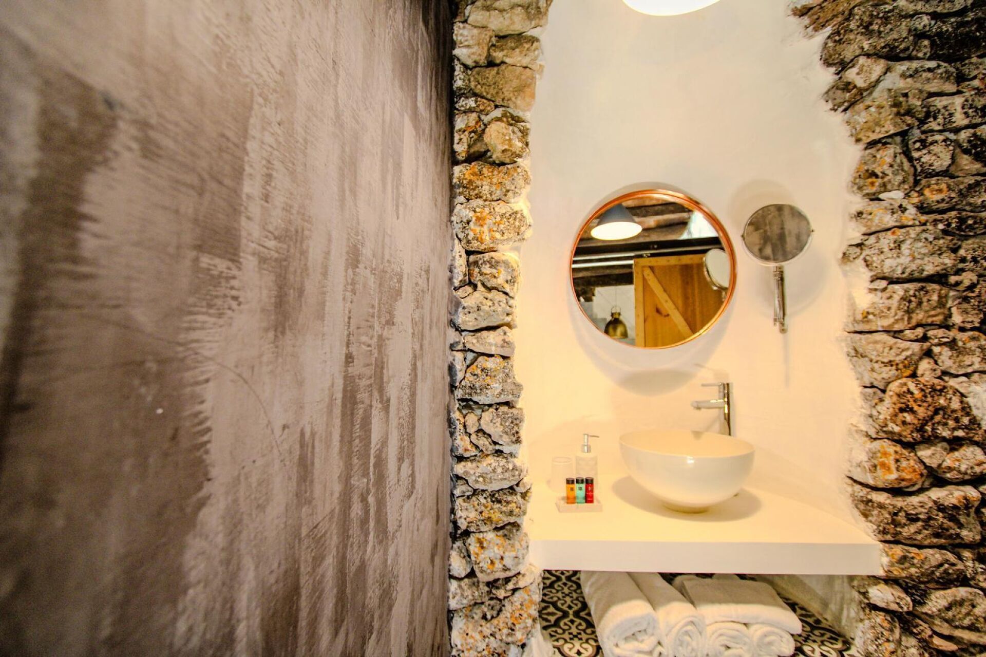 A bathroom with a stone wall, a round mirror, a sink, and a towel rack.