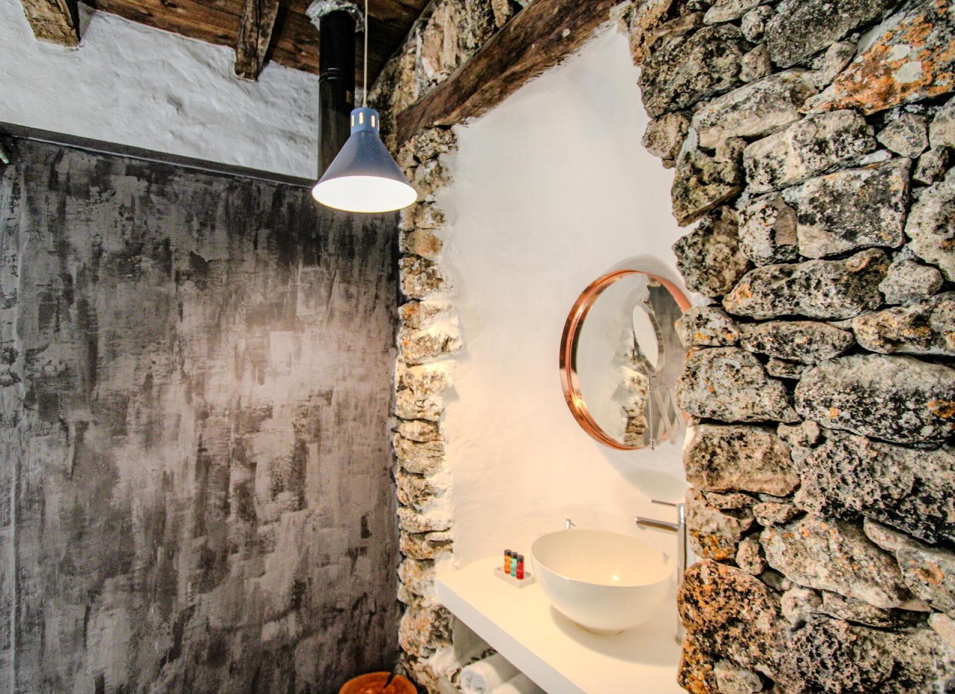 A bathroom with a stone wall, a white sink, a round mirror, and a pendant light.