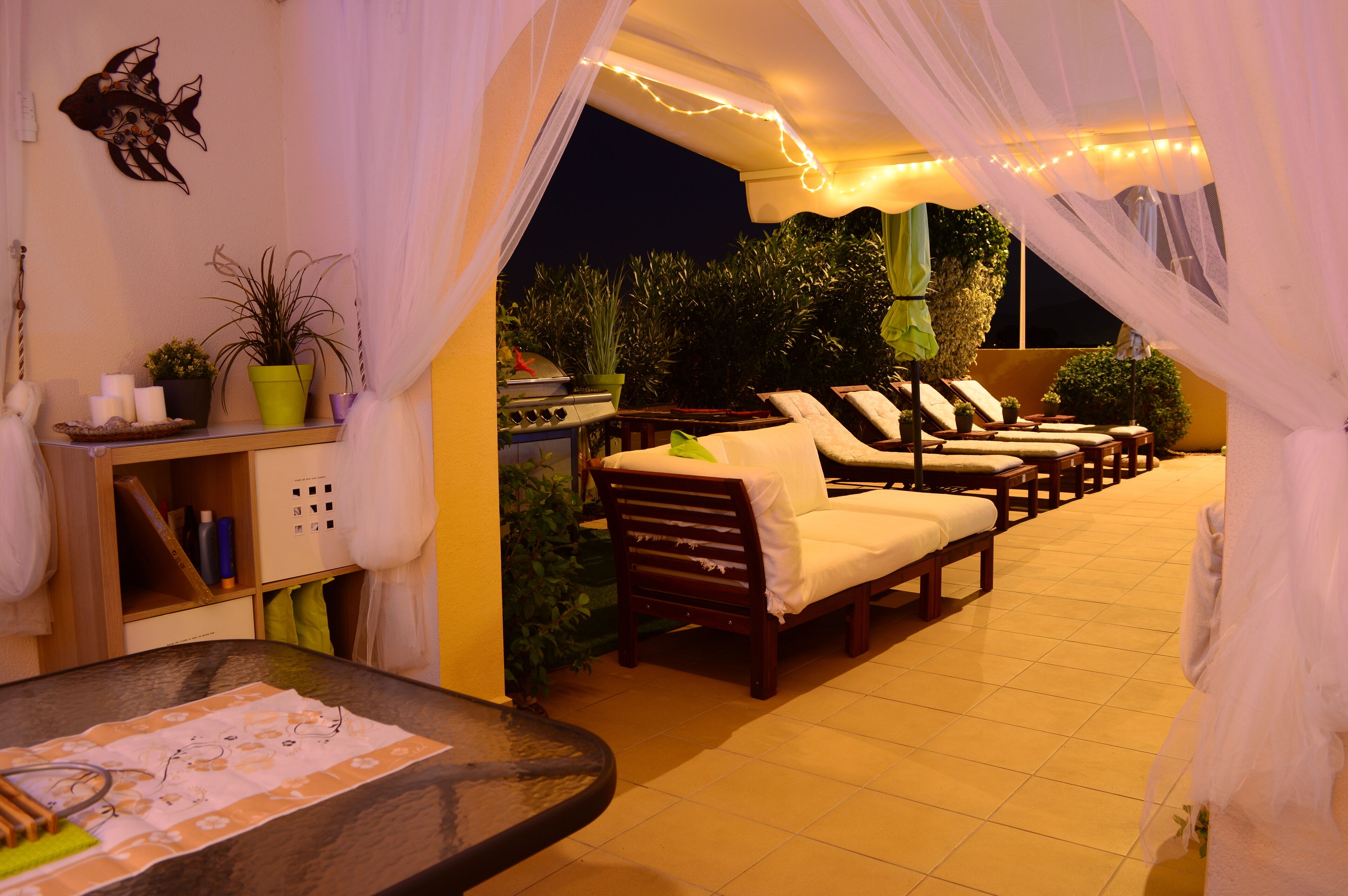 An outdoor seating area with cushioned chairs and a table, surrounded by potted plants and a decorative wall piece.