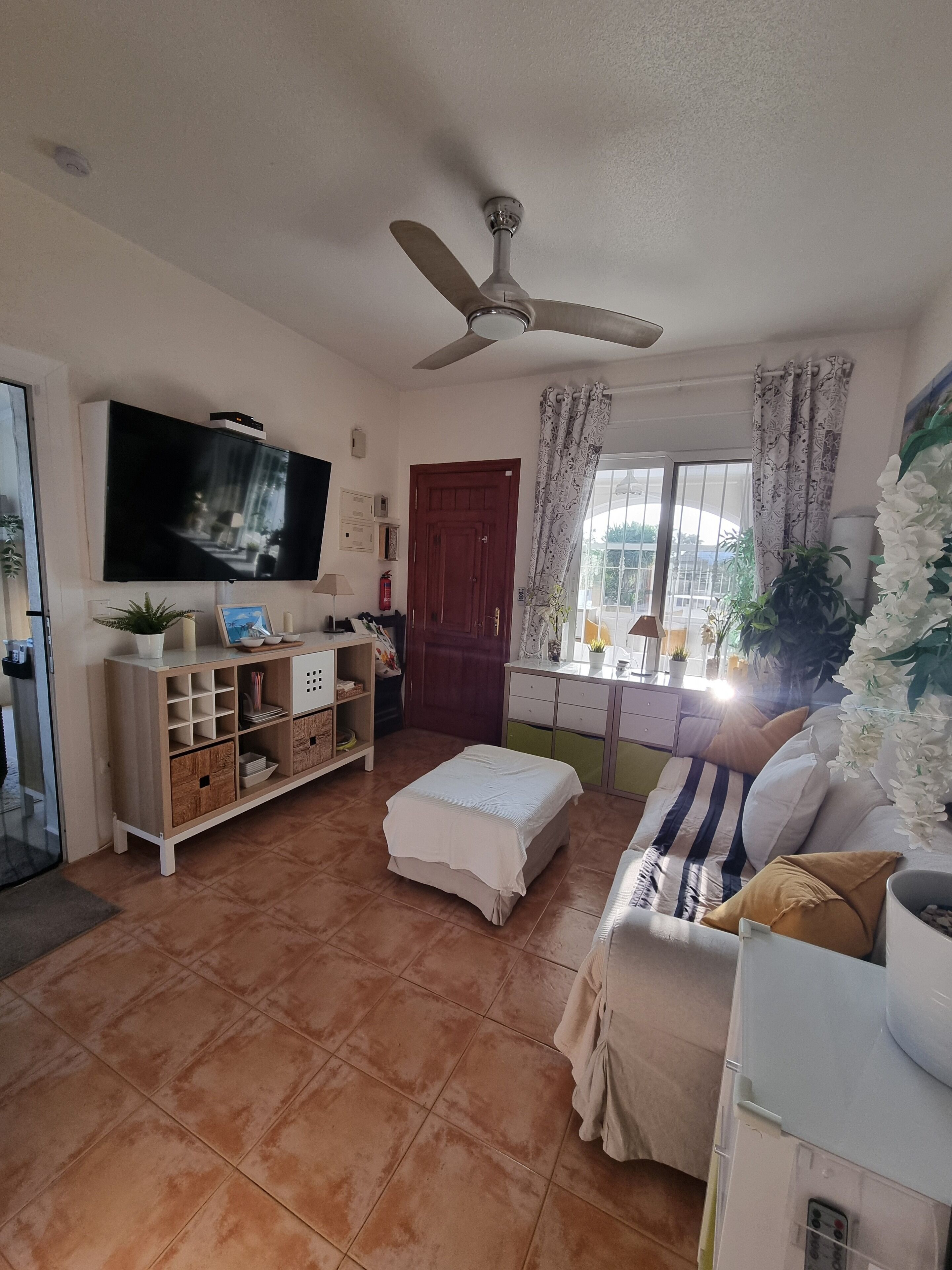 Een woonkamer met een flatscreen-televisie, een plafondventilator, een bank, een salontafel en een raam met gordijnen.