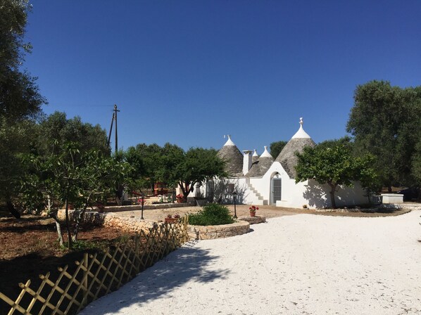 A white building with multiple conical roofs, surrounded by trees and a sandy area.