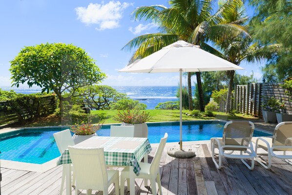 Une piscine avec vue sur l’océan, entourée de palmiers et dotée d’une terrasse en bois avec des chaises et une table.