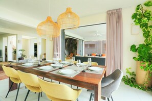 Un coin repas avec une table en bois dressée pour six personnes, des chaises jaunes et grises, et des lampes en osier suspendues au plafond.