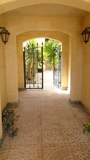 Une entrée en arc surmontée d’un portail en fer forgé, un sol carrelé et une vue sur un jardin planté de palmiers.