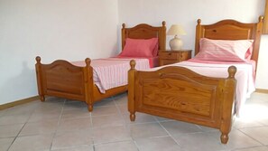 Deux lits en bois avec des têtes de lit, une table de chevet avec une lampe et un sol carrelé.