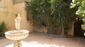 Une statue orne une fontaine située dans une cour, avec un mur de pierre et quelques plantes.