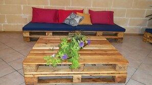 Une table basse rustique en bois, agrémentée d’une plante en pot et de quelques fleurs, est placée devant un canapé bleu orné de coussins rouges et à motifs.