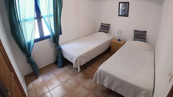 Una habitación pequeña y limpia con dos camas individuales, una mesita de noche, una lámpara y una ventana con cortinas.