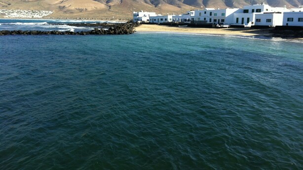 A coastal town with white buildings, a rocky breakwater, and mountains in the background.