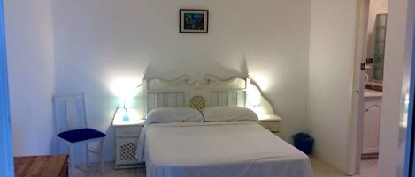 Un dormitorio con cama, mesita de noche, silla y una puerta que da a un baño.