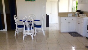 Een eetgedeelte met witte stoelen en een tafel, een keuken met witte kasten, en een deur naar een andere kamer.