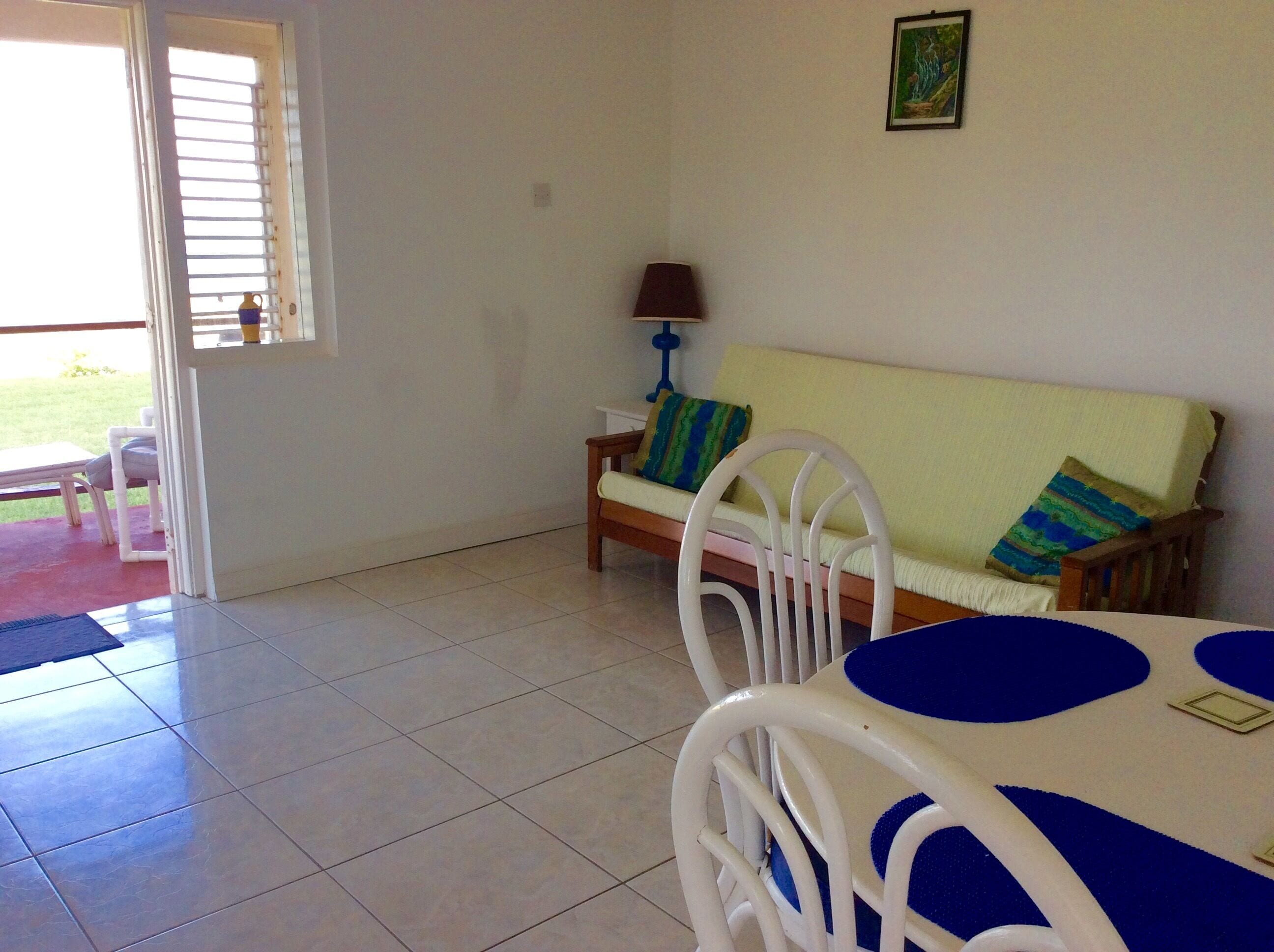 A room with a white sofa, a small table, and a chair. There is a window with blinds, a lamp, and a picture on the wall.