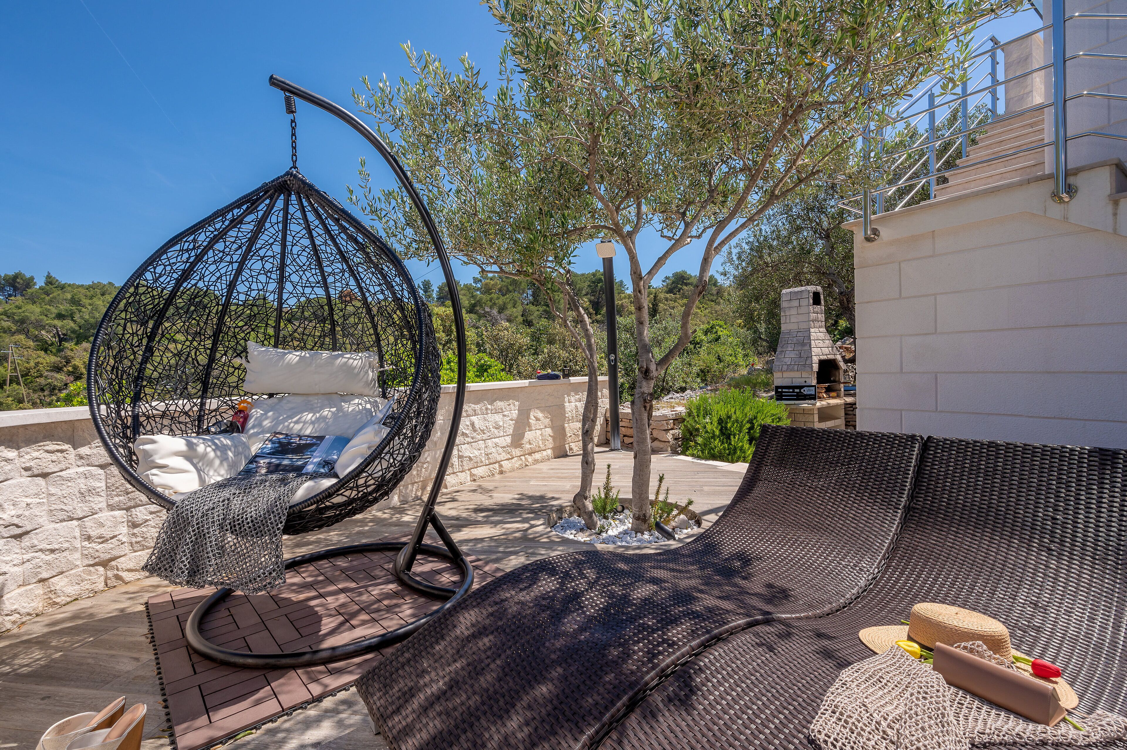 A wicker lounge chair, a hanging egg chair, and a small table with a hat and sunglasses on a patio with a stone wall and trees.