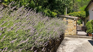 Un muro de piedra con plantas de lavanda, un edificio de piedra con techo de tejas y un cielo azul despejado.