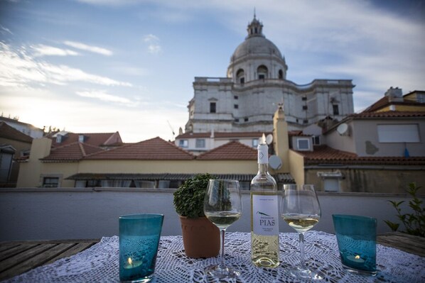 A table with a bottle of white wine, two glasses, a potted plant, and a candle on a lace tablecloth, set against a backdrop of a historic building with a dome.