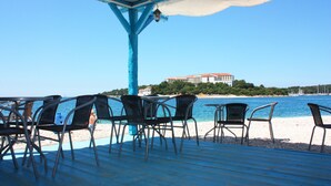 A beachside patio with metal chairs and tables, overlooking a clear blue sea and a building on an island.