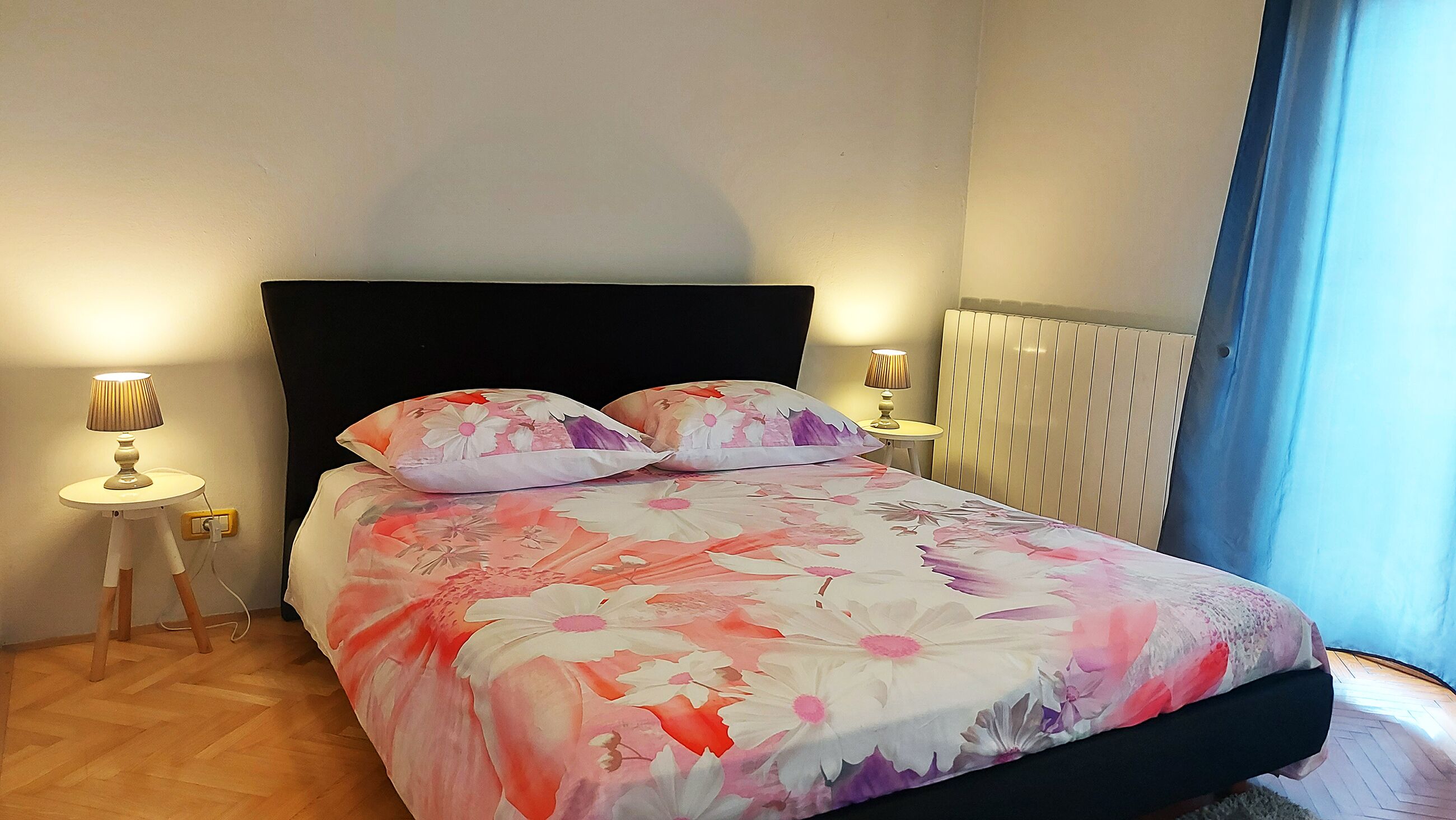 A bedroom with a floral bedspread, two bedside lamps, a radiator, and a view of the outdoors.