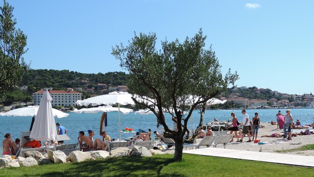 A beach with people sunbathing, umbrellas, and a tree in the foreground. - Authentic Dalmatian Getaway • Pool & free parking (Trogir)