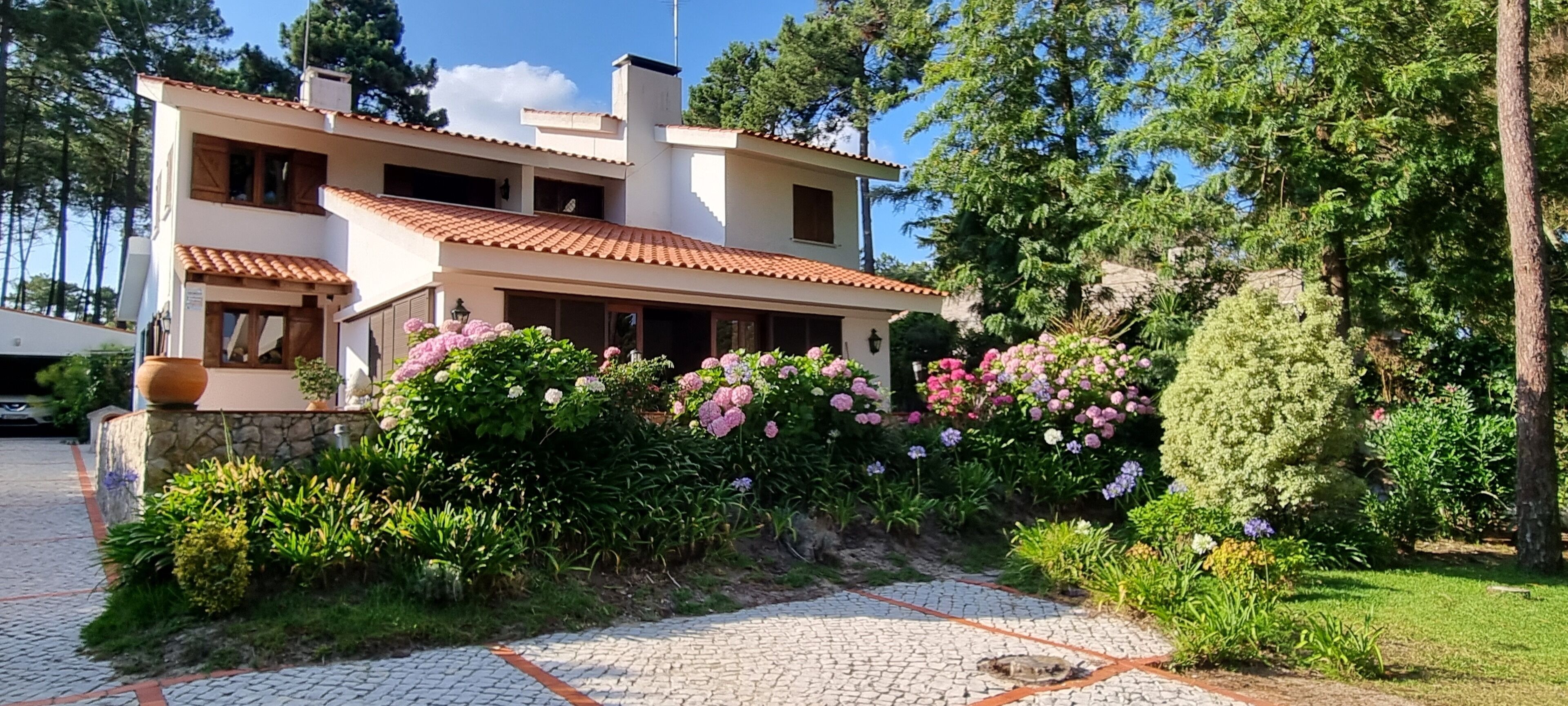 A two-story house with a tiled roof, surrounded by lush greenery and flowering plants.