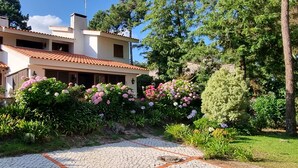 Une maison à deux étages, dotée d’un toit en tuiles, entourée d’une végétation luxuriante et de plantes en fleurs.