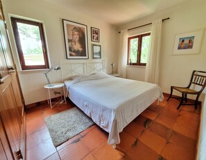 Une chambre à coucher avec un lit, une chaise, une table de chevet, une lampe et une fenêtre avec des rideaux.