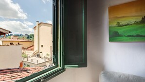 Una habitación con ventana que da a un paisaje urbano, un cuadro en la pared y un sofá con un cojín.