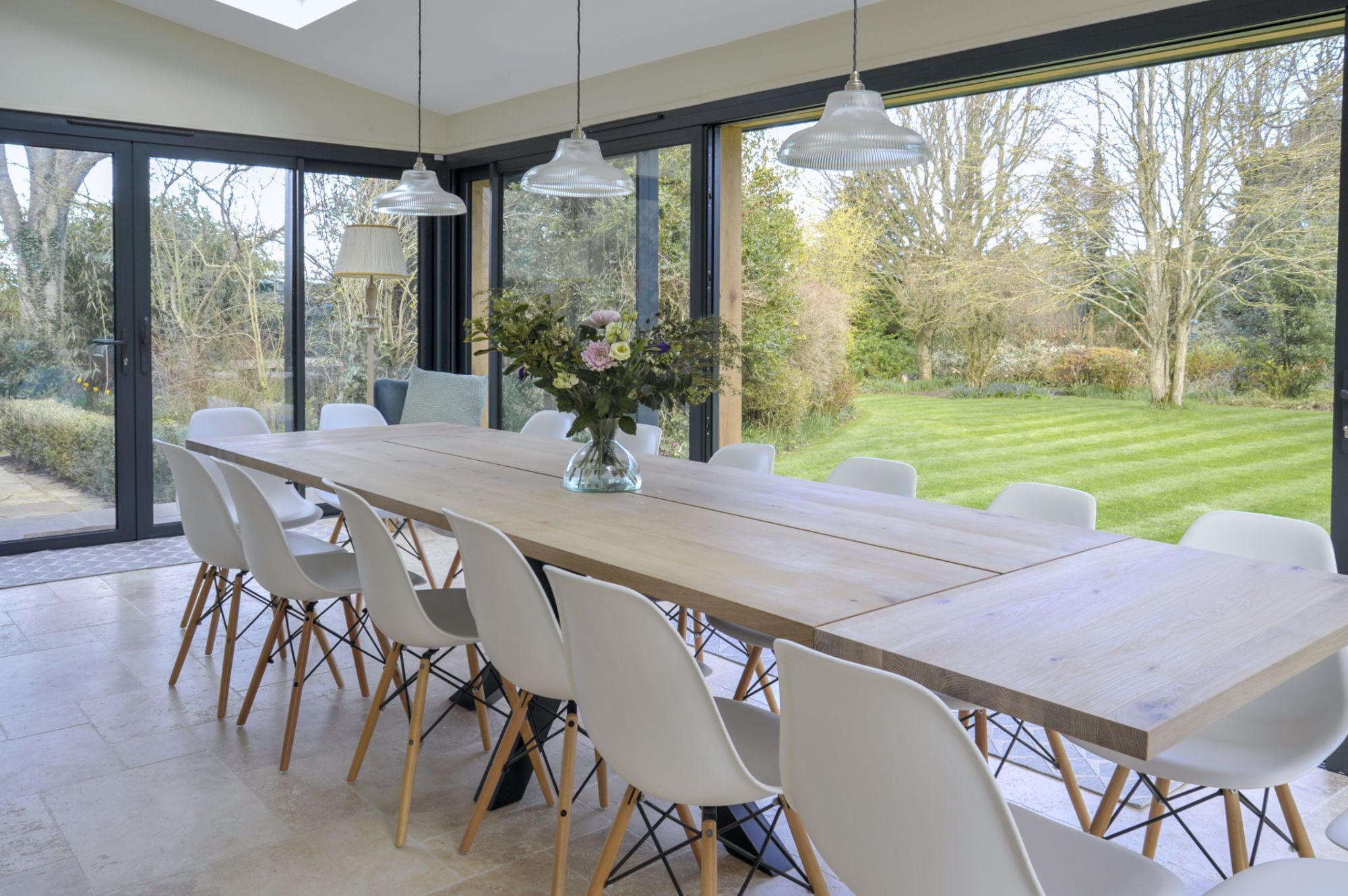 A modern dining area with a long wooden table, white chairs, and a vase of flowers.
