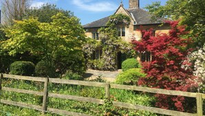 A traditional stone house with a steeply pitched roof, surrounded by lush greenery and a wooden fence.
