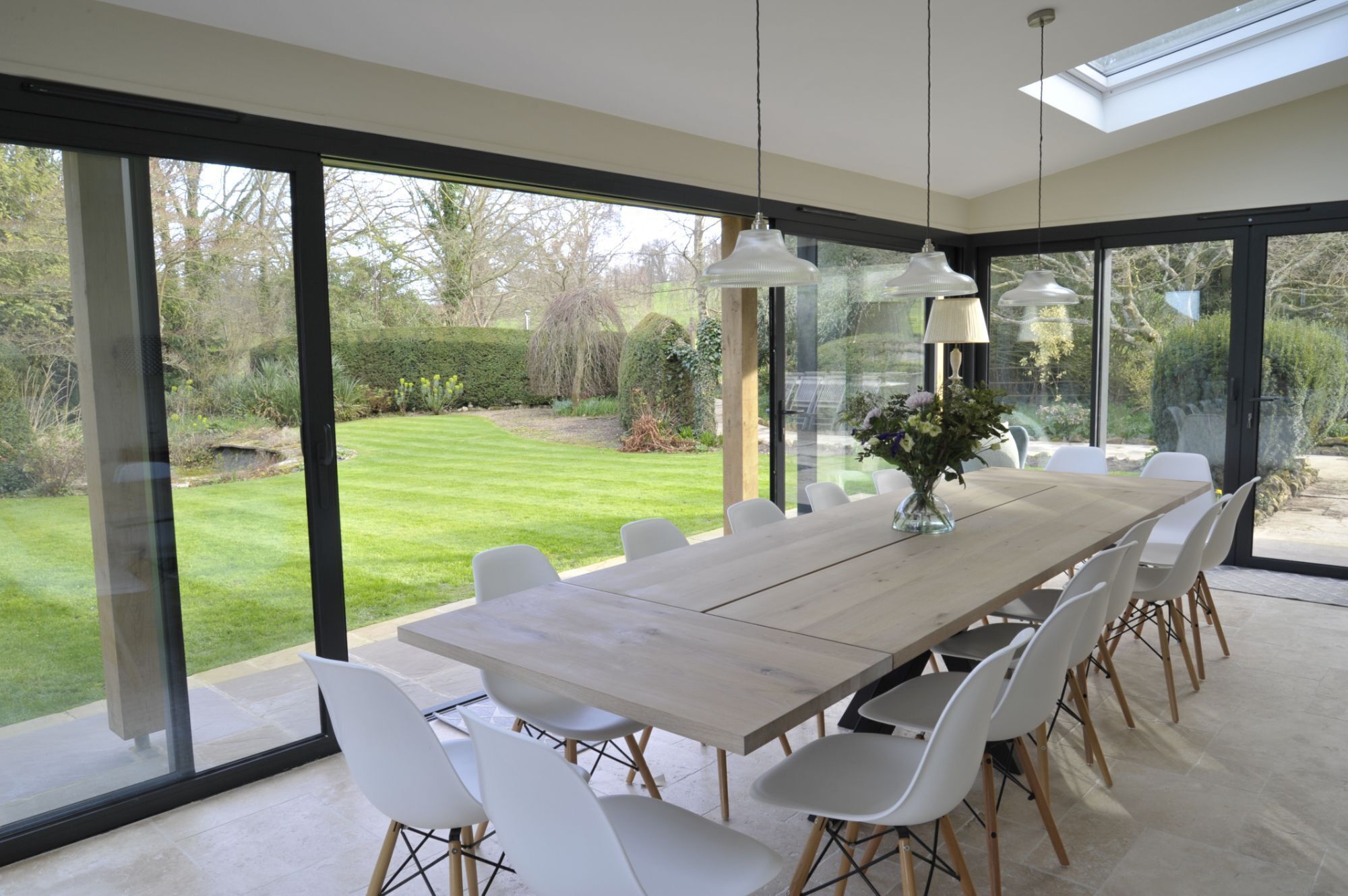 Un espace de restauration moderne avec une grande table en bois et des chaises blanches, ainsi que de grandes fenêtres donnant sur un jardin.