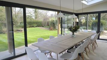 Un espace de restauration moderne avec une grande table en bois et des chaises blanches, ainsi que de grandes fenêtres donnant sur un jardin.