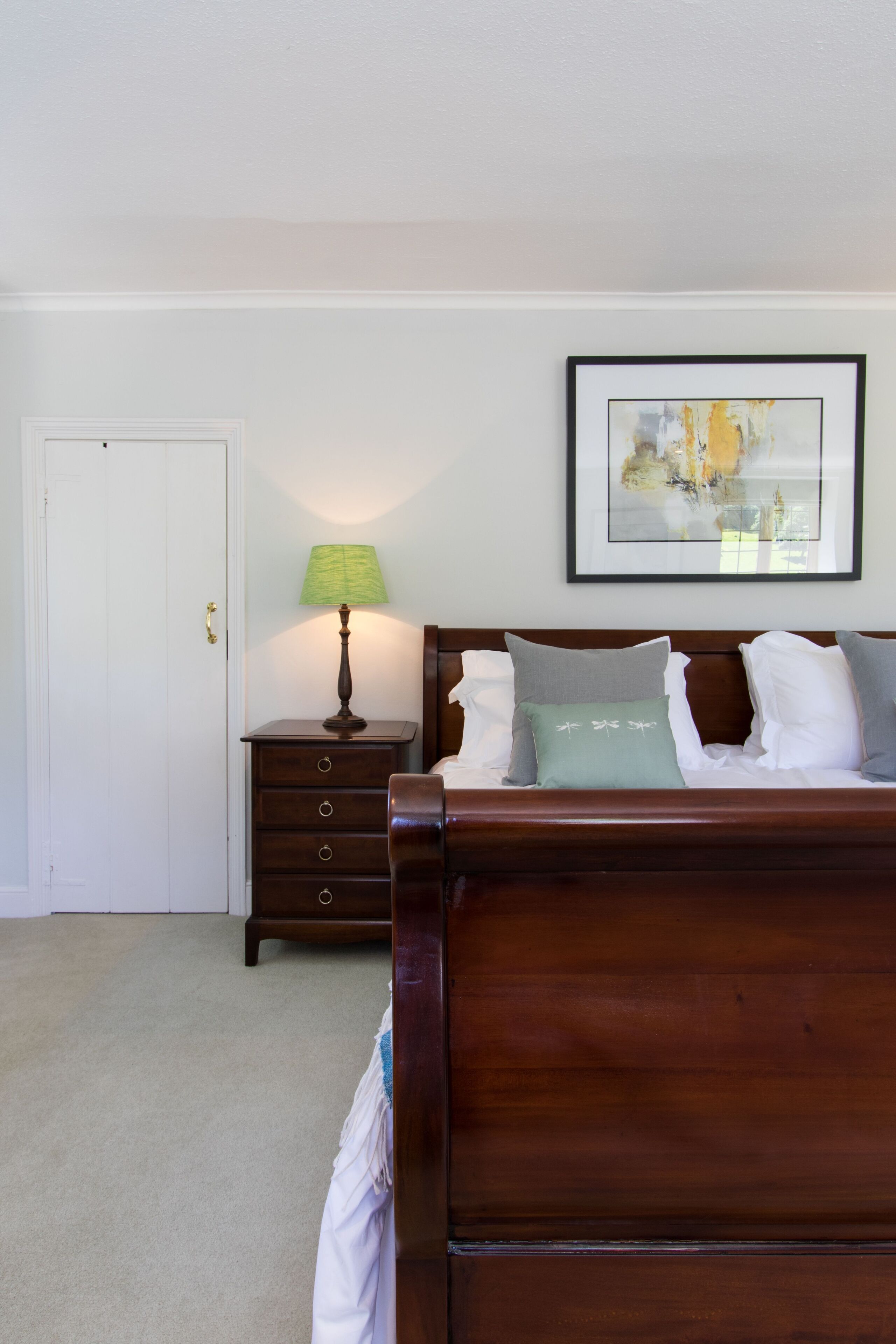 A bedroom with a wooden bed, a nightstand with a lamp, a framed picture on the wall, and a door.