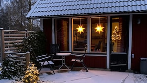 Un rincón al aire libre acogedor con un banco de madera y una mesa, un pequeño árbol de Navidad y un edificio con ventanas amplias y tejado cubierto de nieve.