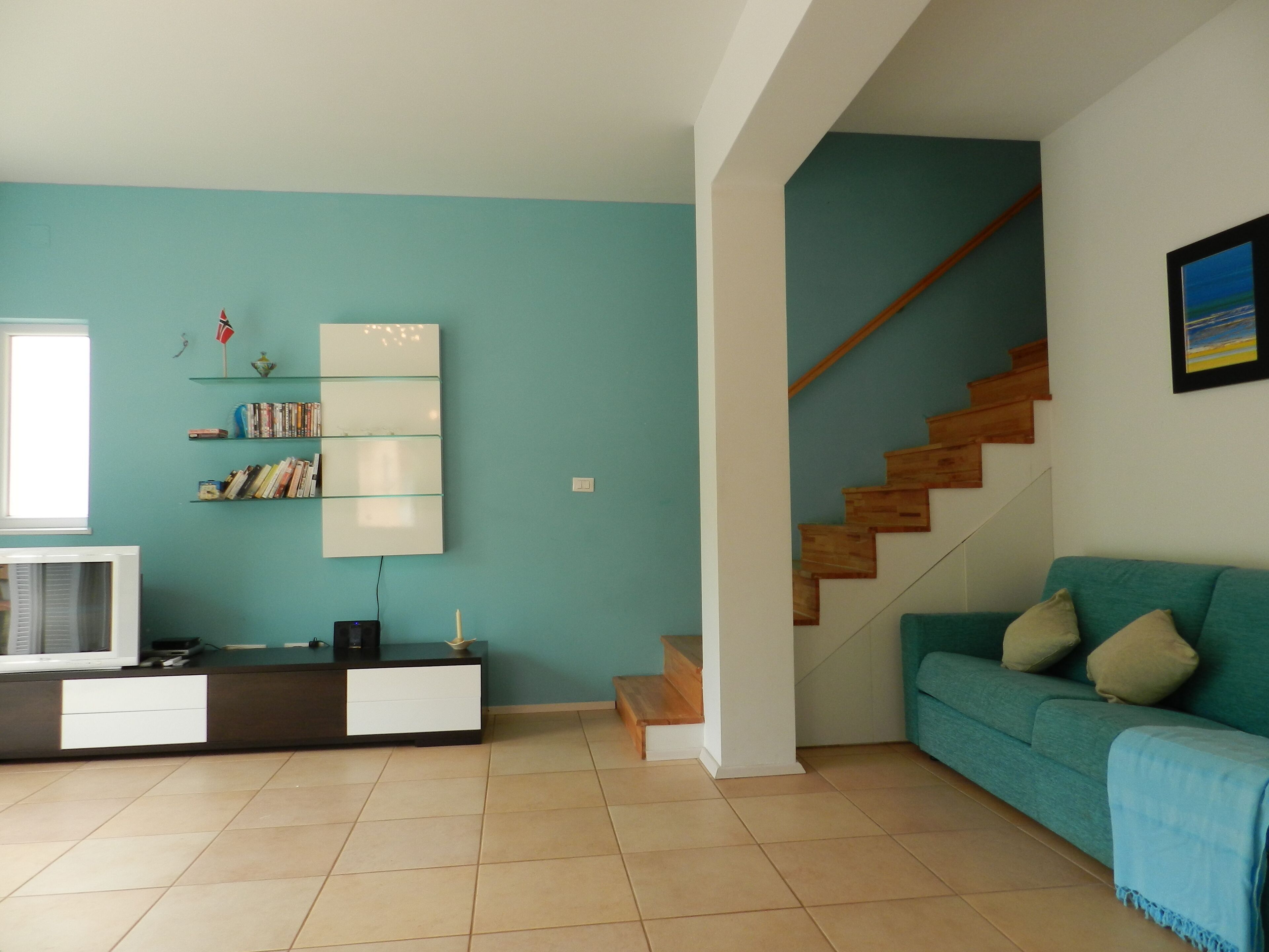 A living room with teal walls, a wooden staircase, a teal sofa, and a white entertainment unit with books.
