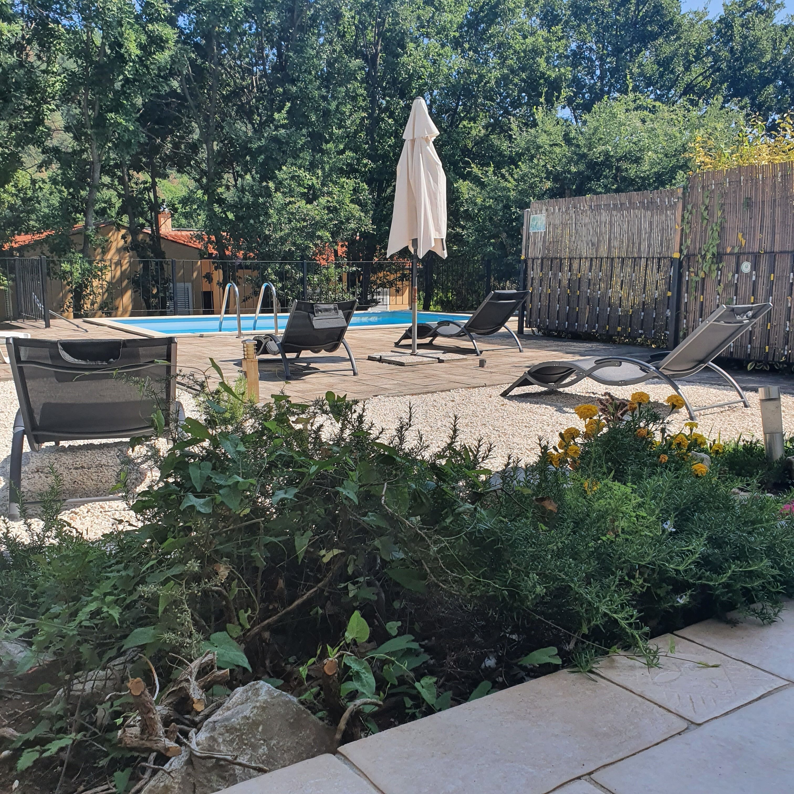 Ein Poolbereich mit Liegestühlen und Sonnenschirm, umgeben von Pflanzen und einem Holzzaun.