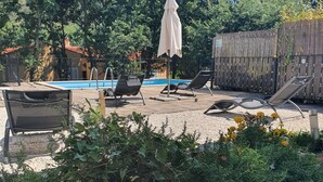A pool area with lounge chairs and a covered umbrella, surrounded by greenery and a wooden fence.
