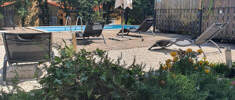 Zona de piscina con sillas de descanso y una sombrilla, rodeada de follaje y una valla de madera.
