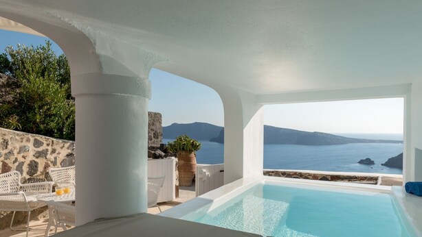 A pool area with a view of the sea and mountains, white furniture, and a stone wall.