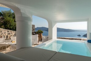 A pool area with a view of the sea and mountains, white furniture, and a stone wall.